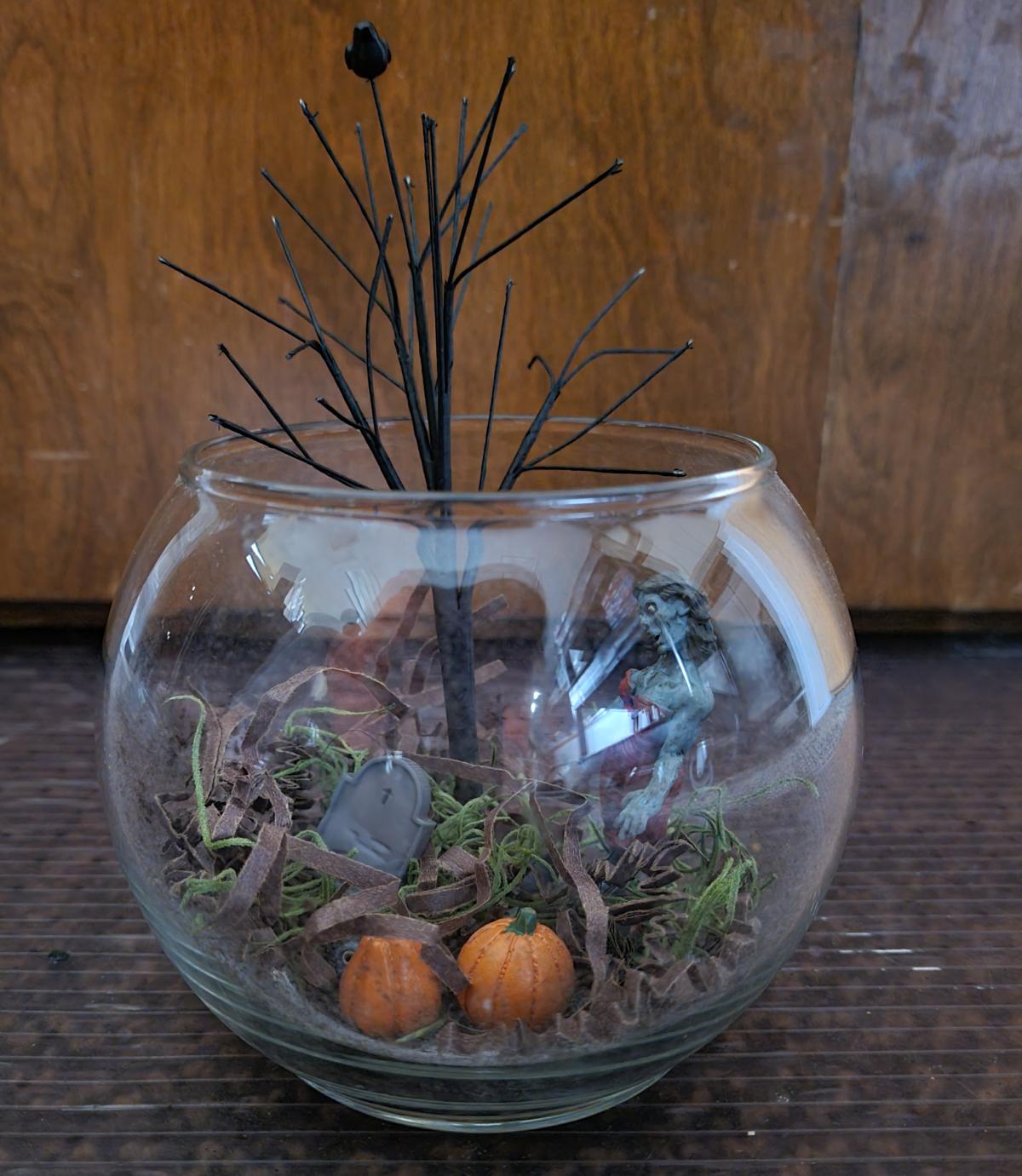 A glass bowl with a zombie, pumpkins and a dead tree inside it.